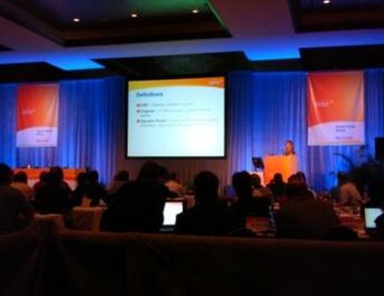 A conference room with a large screen in the center. The screen displays a presentation slide with text that reads "Solutions". On either side of the screen there are two banners with orange and white text. The room is decorated with blue curtains and there are people sitting at tables with laptops in front of them. The people in the room appear to be engaged in the presentation. The overall atmosphere of the room is professional and organized.