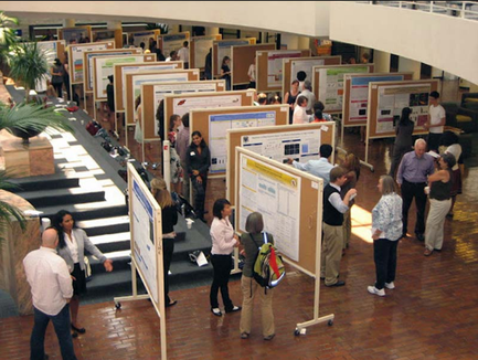 A large group of people gathered in a large room with a high ceiling and a staircase in the background. The room appears to be an art gallery or exhibition space as there are several large posters and posters on display on easels. The people are of different ages and genders and they are all looking at the posters with interest. Some of them are standing in front of the posters while others are walking around the room. There are plants and other artworks scattered throughout the space. The overall atmosphere of the image is lively and vibrant.