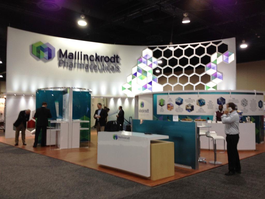 A trade show booth with a white and blue color scheme. The booth has a large banner that reads "Mallinckrodt Pharmaceuticals" in bold colorful letters. The banner also has a geometric design with different shades of blue green and purple. There are several people standing in front of the booth some of them are looking at the display. On the right side of the image there is a desk with a computer and a few other items on it. In the background there are other booths and people walking around. The overall atmosphere of the exhibition hall is modern and professional.